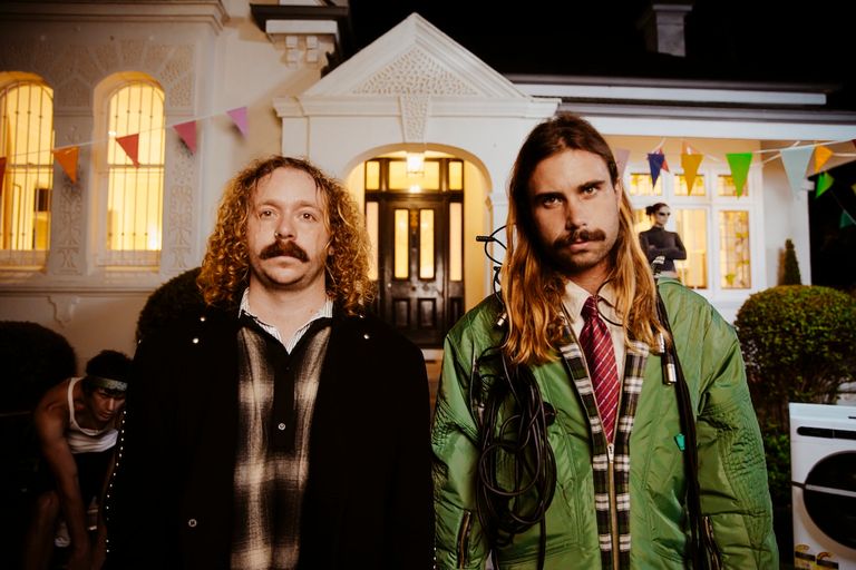 Australian music duo Hellcat Speedracer standing outside a suburban house with colorful party bunting at night, looking directly at the camera.